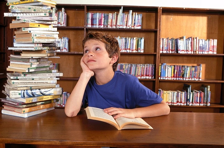 ילד בספרייה, ילד מיואש מללמוד, ילד מביט בערימת ספרים על שולחן מבספריה כשהוא קורא בספר
