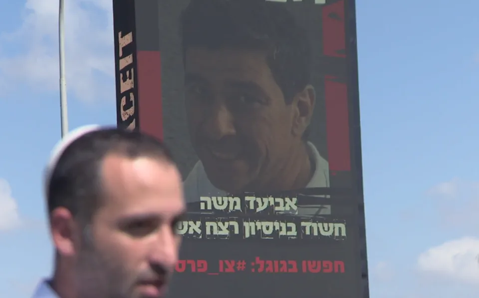 אחיה של שירה איסקוב ברקע שלט החוצות עם תמונתו של החשוד בניסיון לרצח - אביעד משה